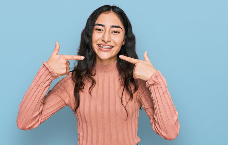 Girl in turtleneck smiling and pointing at her toothy grin with braces using both hands.